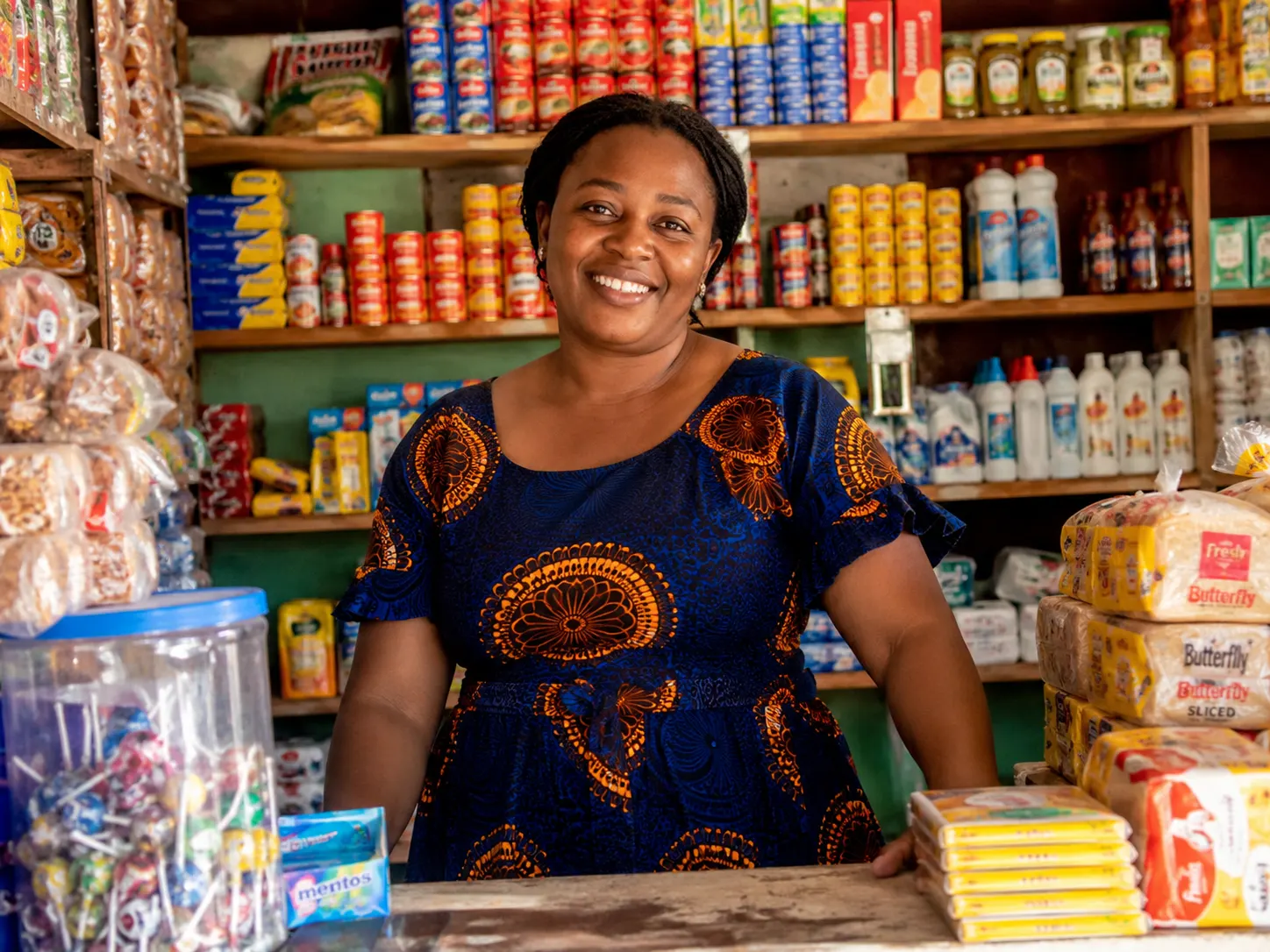 Smiling African shop owner in a patterned dress behind a counter stocked with bread, canned goods and household items